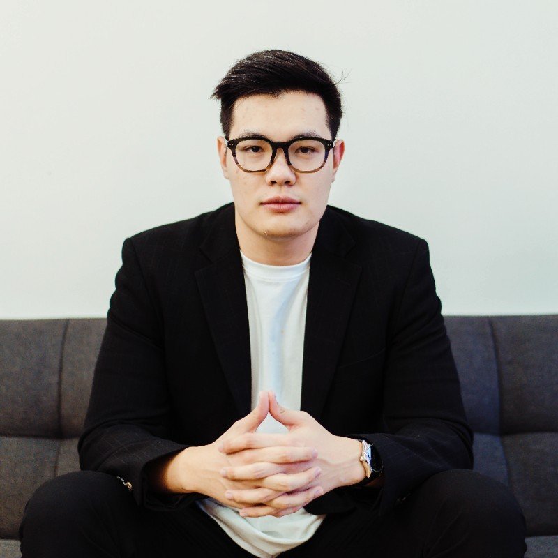 Max Do wearing glasses and a black blazer, seated on a sofa